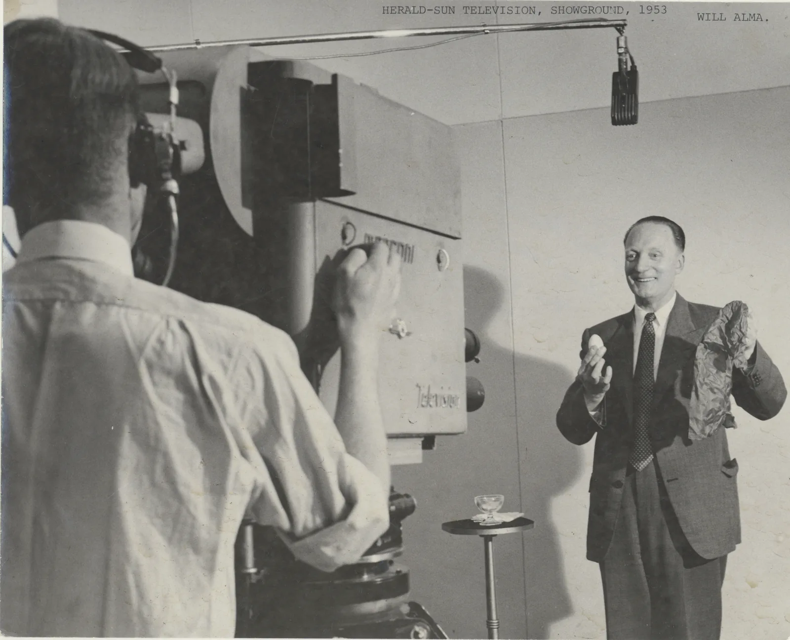 A smiling magician in a double-breasted suit holds up an egg and a cloth before a large studio camera lettered Marconi and Television; the cameraman, in headphones, has his back to us, and a boom microphone hangs overhead.