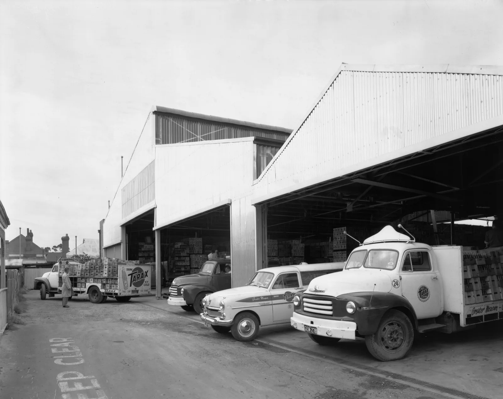Delivery trucks and a van pull out from under a saw-tooth loading-bay roof stacked with crates; the round Tarax badge is on a truck door, and Keep Clear is painted on the road.