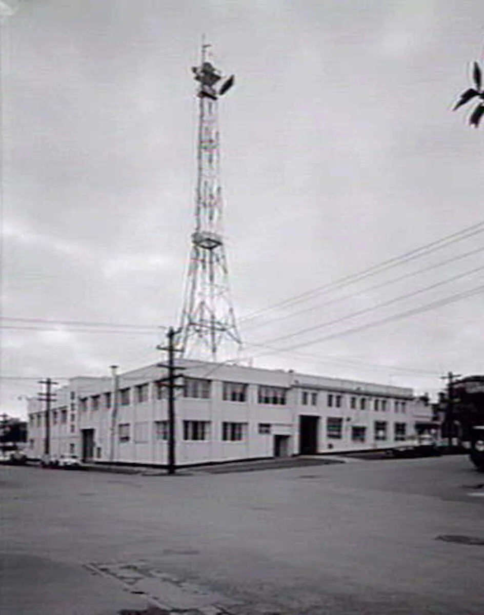 A plain two-storey white studio building on a street corner, a lattice tower with a dish rising behind it, power poles and wires against an overcast sky.
